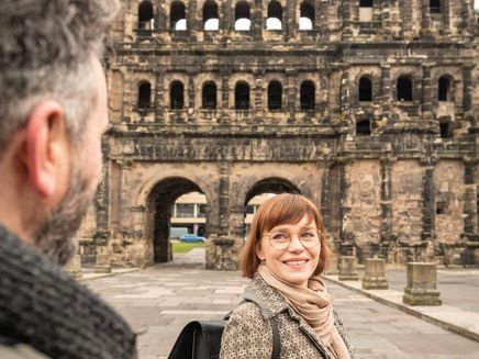 Trier, Porta Nigra © D. Ketz / MLT GmbH Frau mit Mantel und schwarzem Rucksack vor Porta Nigra blickt über Schulter zu Mann
