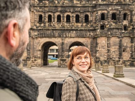 Trier, Porta Nigra © D. Ketz / MLT GmbH Frau mit Mantel und schwarzem Rucksack vor Porta Nigra blickt über Schulter zu Mann