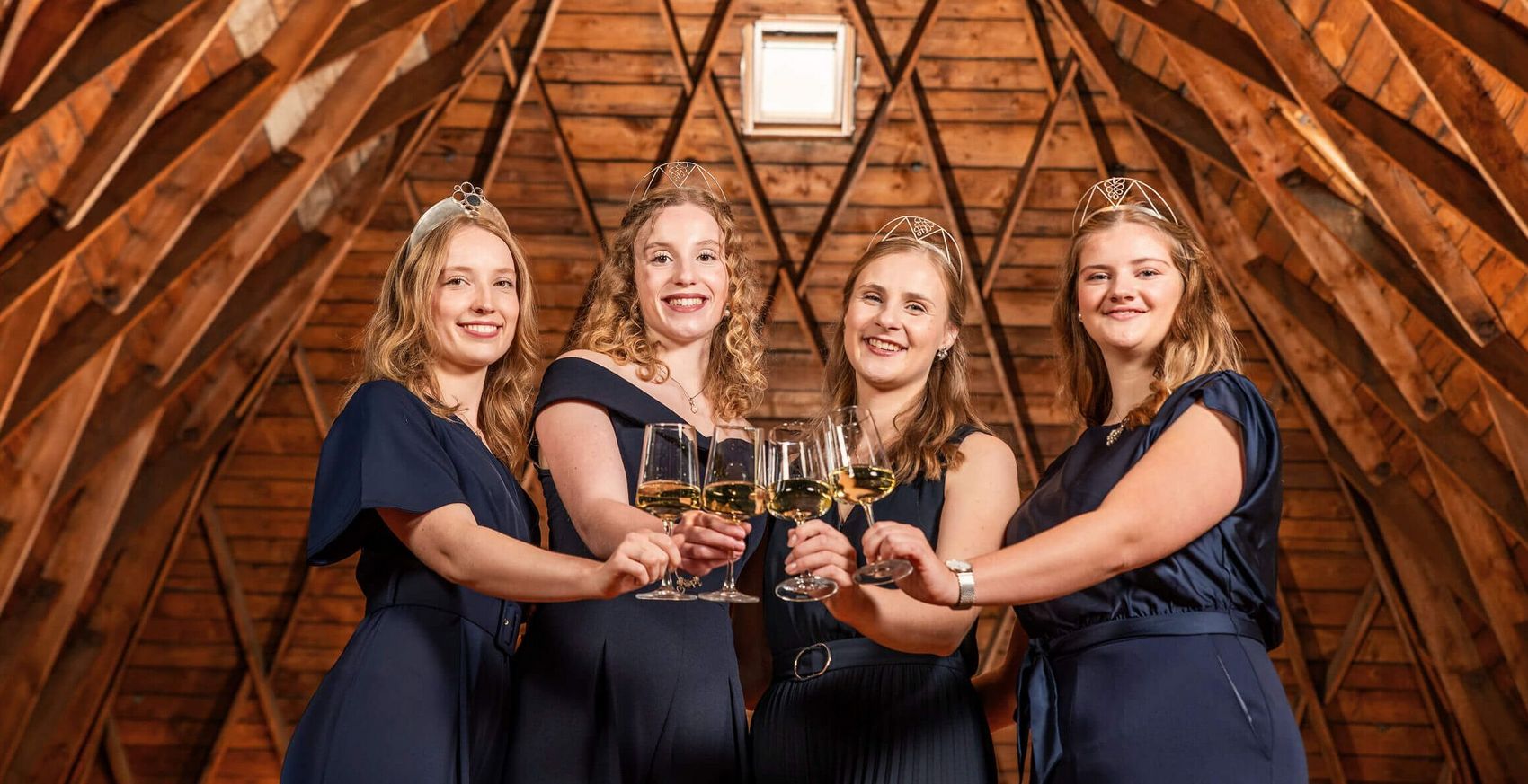 Moselweinhoheiten 2024/2025 © Chris Marmann/Moselwein e.V. Vier Frauen mit Kronen in blauen Kleidern schauen in die Kamera und stoßen mit Weingläsern an.