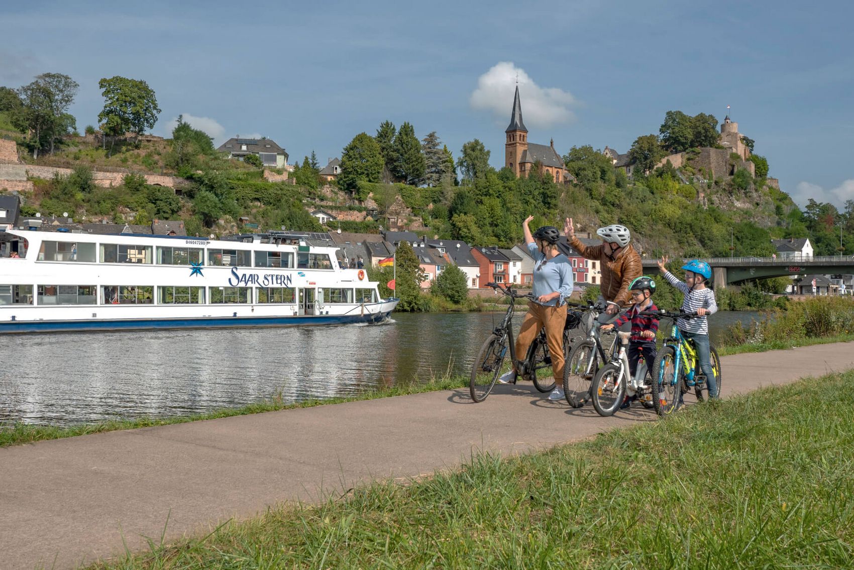 Saar-Radweg bei Saarburg © Hans-Peter Merten Ansicht auf eine Familie bei einer Fahrradtour auf dem Saar-Radweg, die gerade einem vorbeifahrenden Schiff zuwinkt.